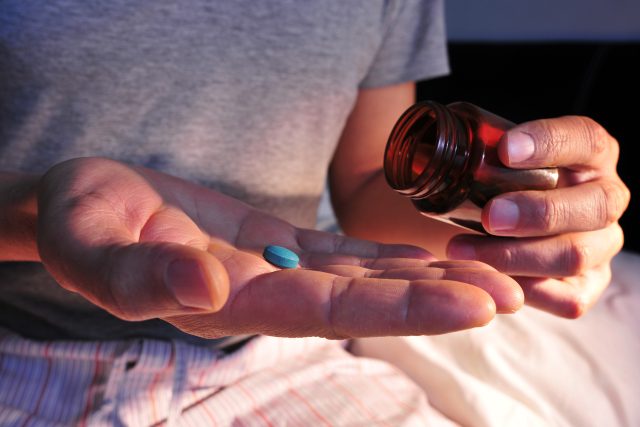 Homem sentado na cama com a mão cheia de comprimidos azuis representando a sildenafila