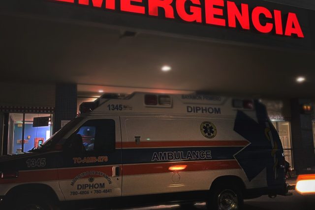 Imagem de hospital com letreiro vermelho de emergência ao fundo e uma ambulância na frente com um paciente buscando causas de dor no pênis