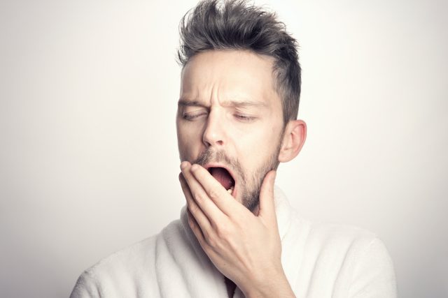 Fundo branco e homem branco com cabelos e barba castanhos bocejando com a mão na boca simbolizando os efeitos do sono na saúde sexual masculina