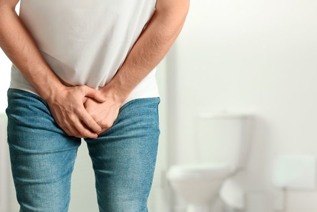 Incontinência urinária e disfunção erétil: Homem branco vestindo camiseta na cor branca e calça jeans, com as duas mãos sobre a calça, na altura do pênis. No fundo da imagem, uma privada.