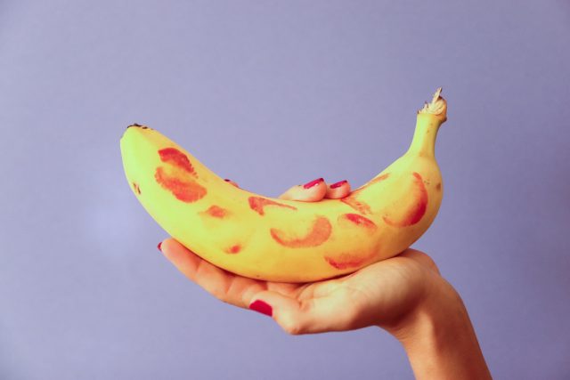 A imagem mostra uma banana com casca coberta de beijos de batom vermelho sobre fundo roxo simulando a sensação de prazer com prótese peniana